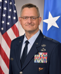 A headshot of Dr. Timothy Grayson in a tan suit with a navy tie infront of an American flag
